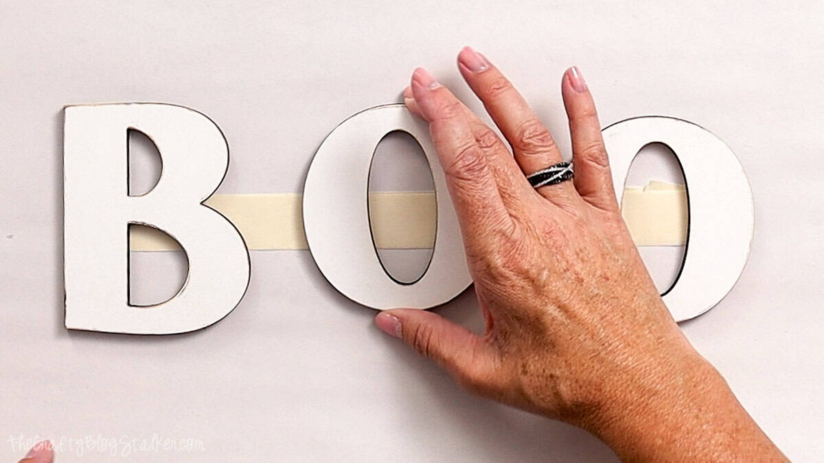 Hand positioning white wooden BOO letters over masking tape to keep them aligned for a Halloween door hanger project.