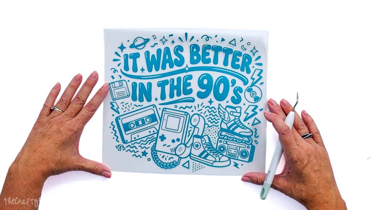 Hands holding a weeded teal puff vinyl 90s design on its clear carrier sheet with a Cricut weeding tool.