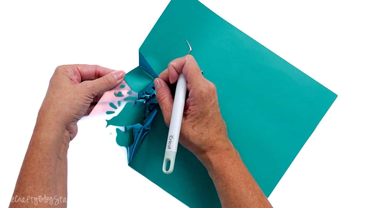 Hands weeding excess teal puff vinyl from a cut design using a Cricut weeding tool.