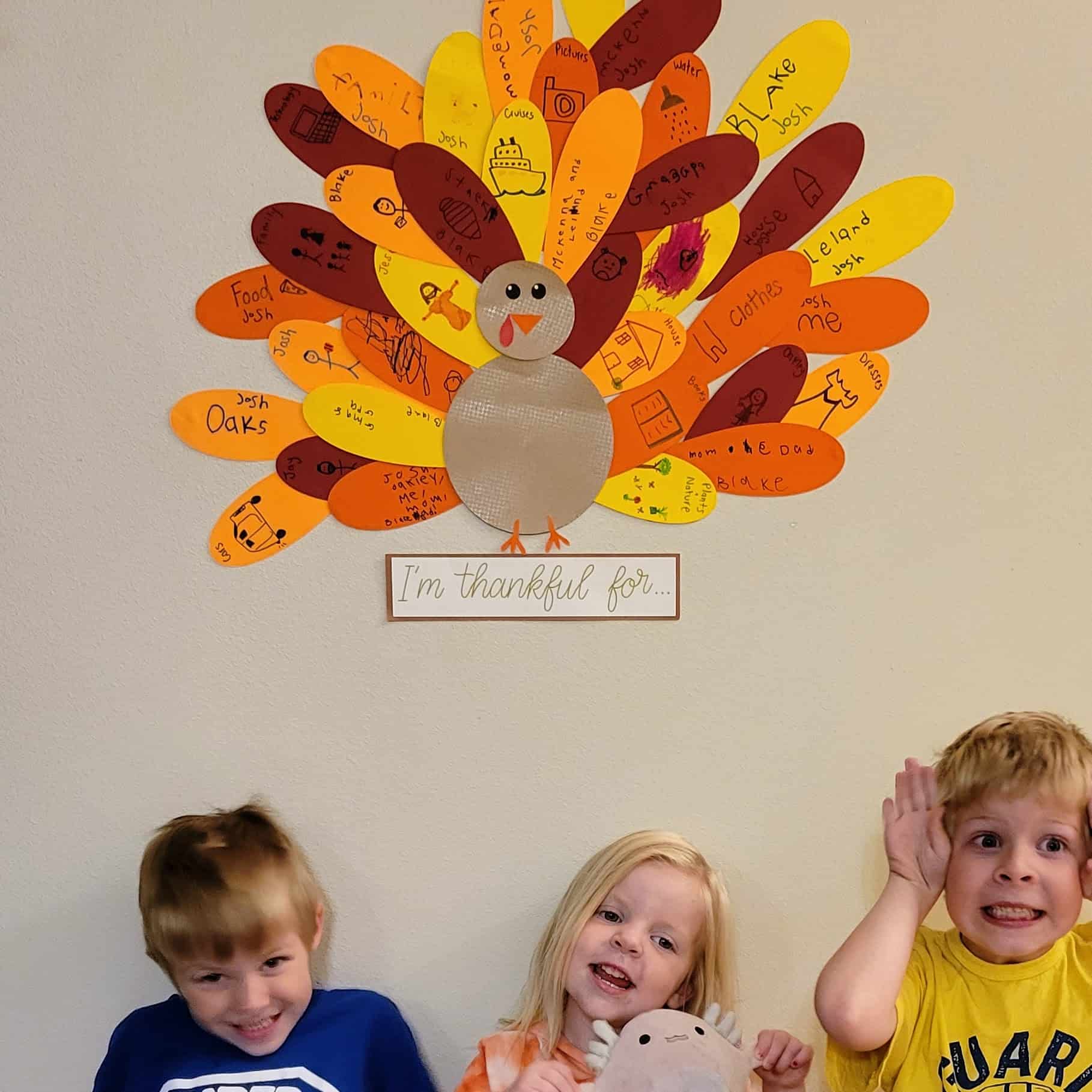 Three children sit beneath a colorful paper turkey gratitude display with feathers showing drawings and handwritten things they are thankful for.