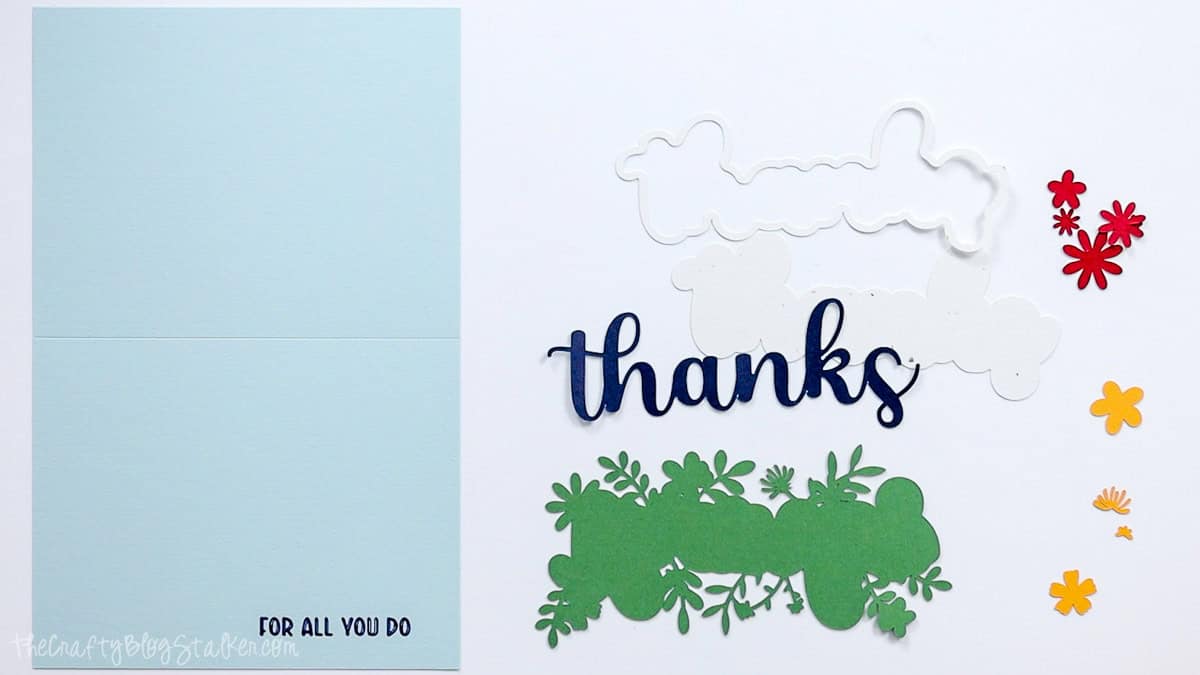 Cut cardstock pieces for a Cricut thank you card, including blue “thanks” lettering, white layers, green foliage, colorful flowers, and card base.
