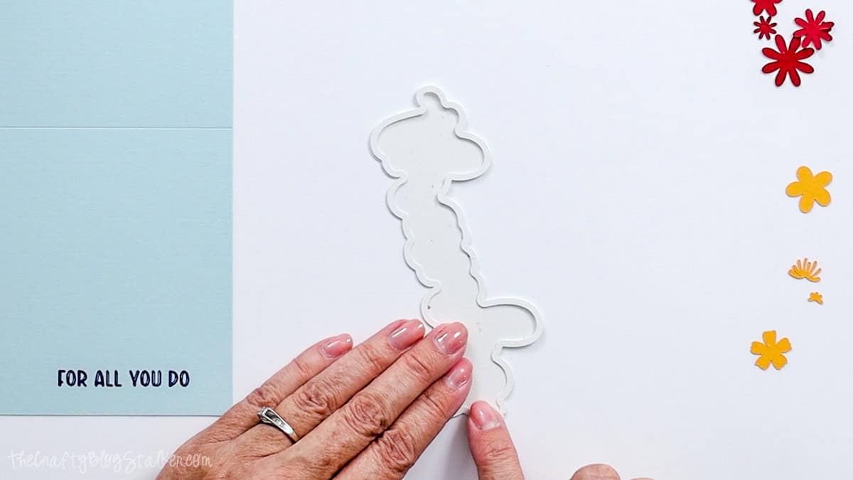 Hands aligning white cardstock frame layers for a Cricut thank you card, with the blue card base and colorful flower pieces nearby.