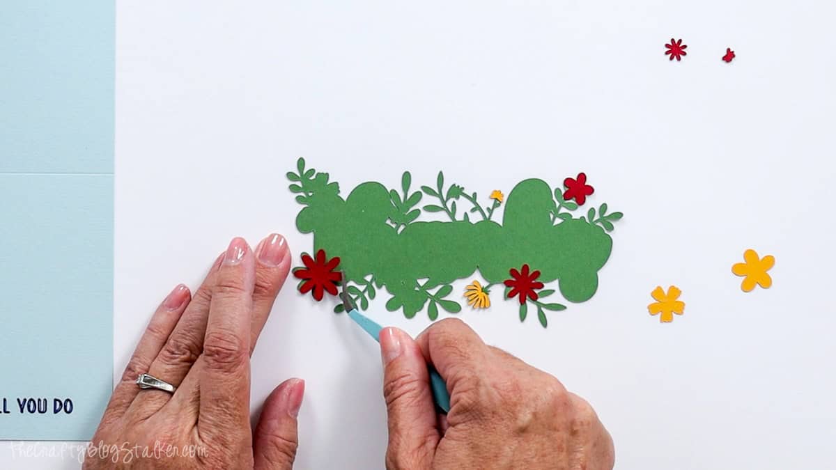 Hands using a precision tool to arrange red and yellow cardstock flowers on green foliage for a Cricut thank you card.