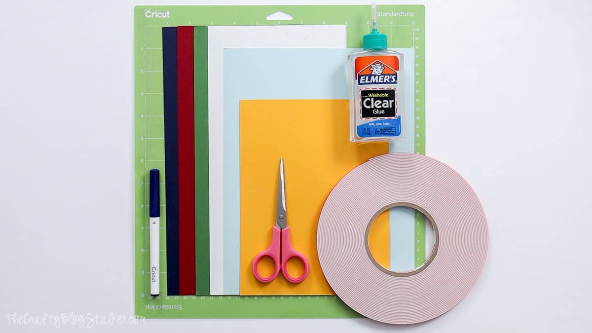 Cricut card-making supplies including colorful cardstock, cutting mat, pen, scissors, clear glue, and foam adhesive tape.