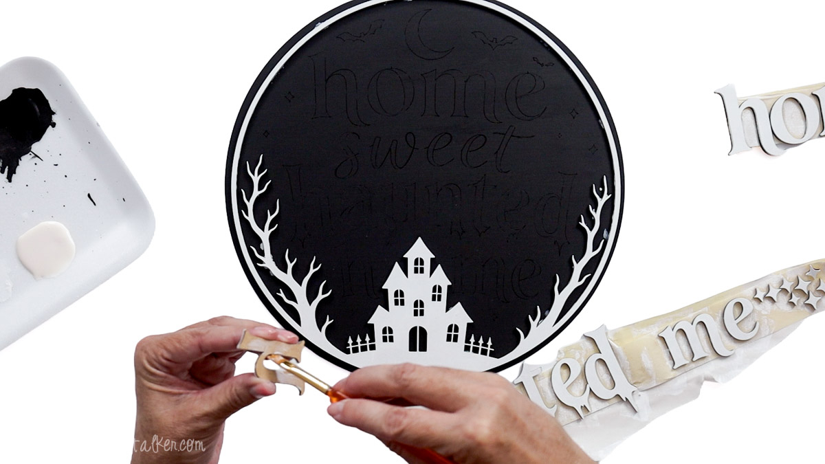 Brushing glue onto white laser-cut lettering for a black Home Sweet Haunted Home sign with a white haunted house and bare trees.
