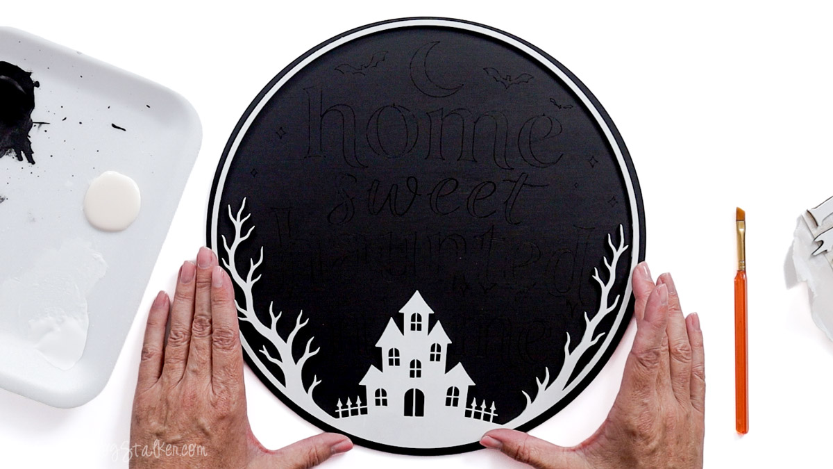 Black circular Home Sweet Haunted Home sign with a white laser-cut haunted house, bare trees, and border being assembled.