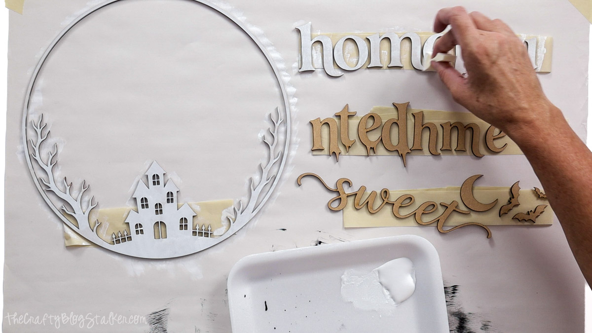 Painting laser-cut wood pieces white for a Home Sweet Haunted Home sign, including spooky lettering, a haunted house, trees, moon, and bats.