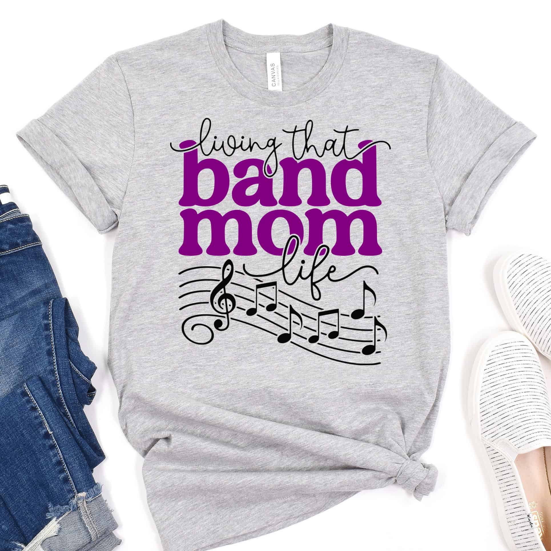 Light gray Band Mom Life T-shirt with purple and black lettering, musical notes, and a treble clef, displayed with jeans and white slip-on shoes.