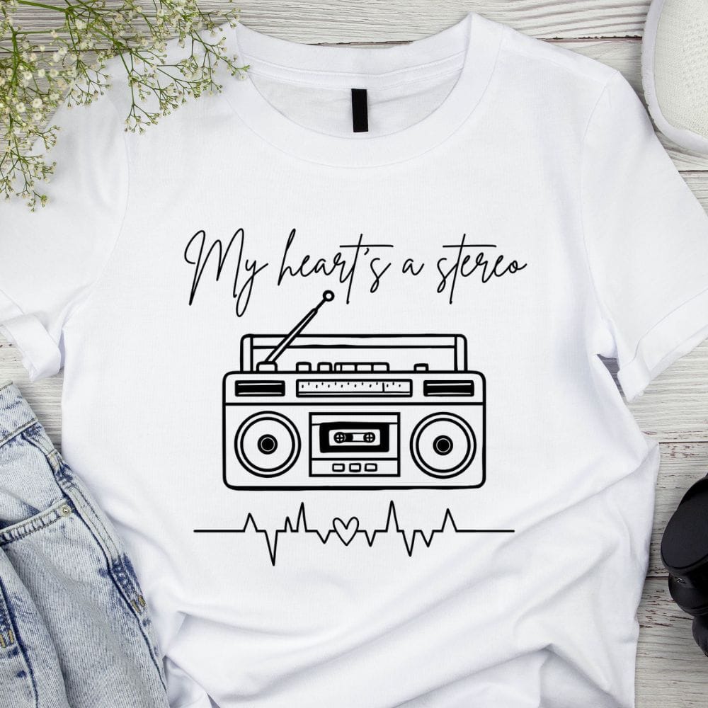 White T-shirt with "My Heart's a Stereo" in black script above a retro boombox and heartbeat line with a heart, displayed beside denim jeans.