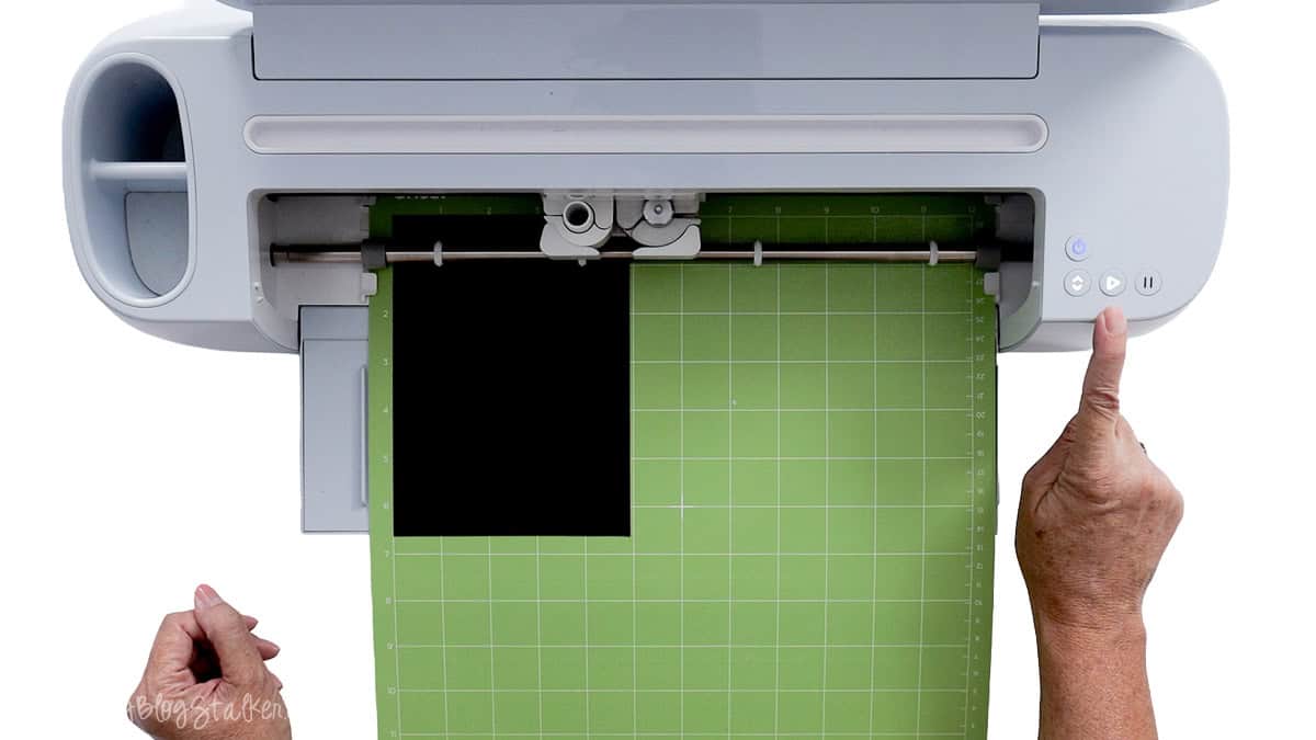 Hand pressing the start button on a Cricut cutting machine with black cardstock attached to a green StandardGrip cutting mat.