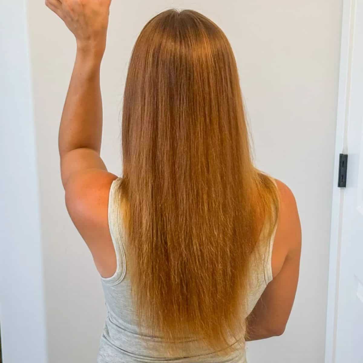Back view of a woman with long, straight, reddish-blonde hair reaching her lower back, wearing a light gray sleeveless top.