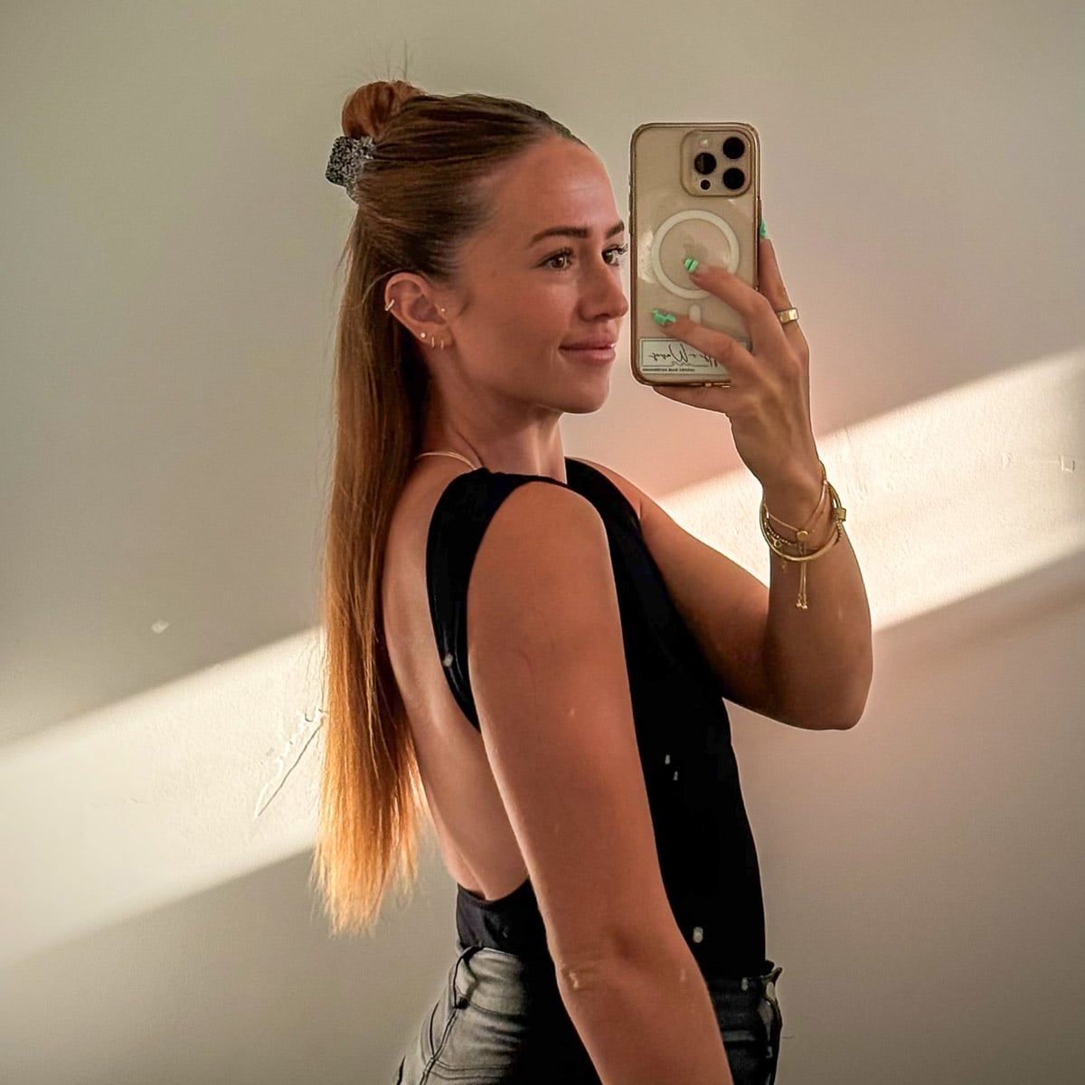 Woman taking a mirror selfie with long, straight reddish-blonde hair styled half-up in a small bun, wearing a black sleeveless top.