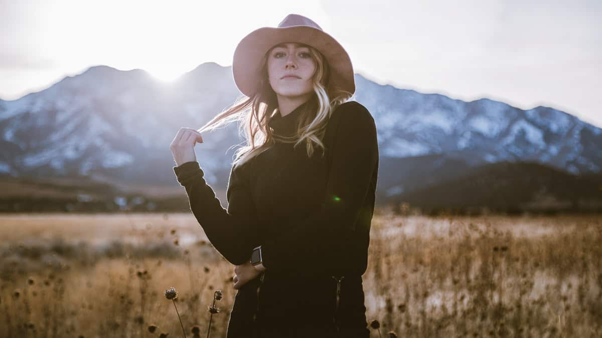 Woman with shoulder-length wavy blonde hair wearing a wide-brimmed hat and black outfit in a grassy field with snow-covered mountains behind her.