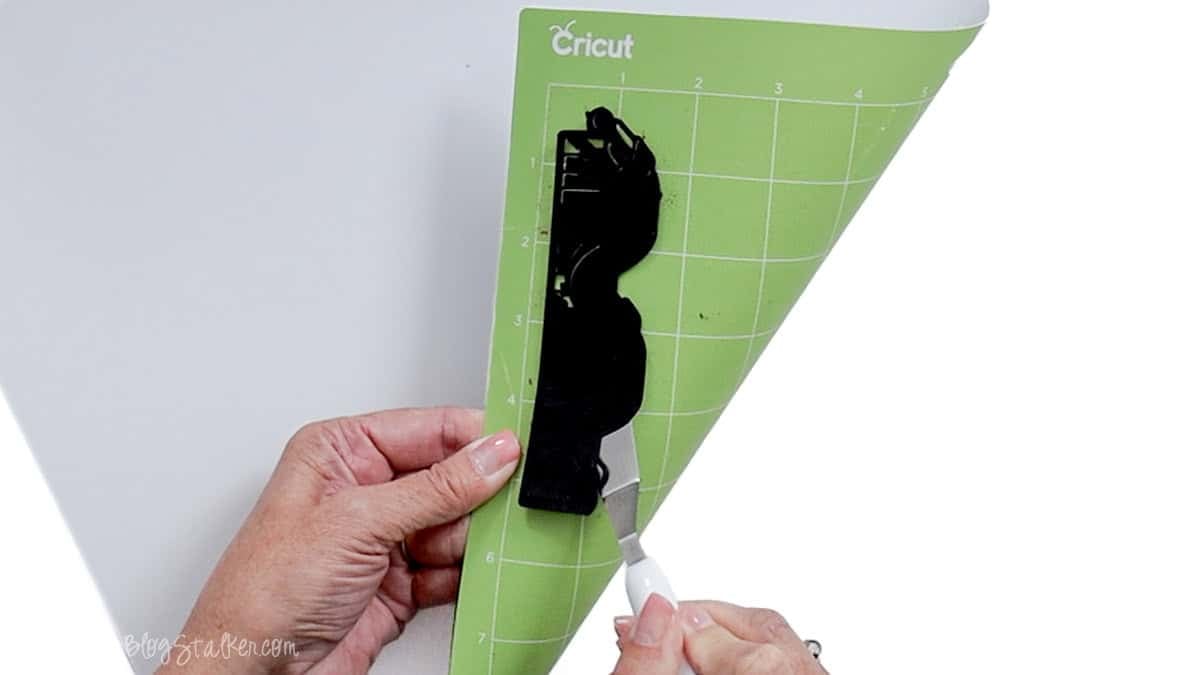 Hands removing a black cardstock piano bookmark cutout from a green Cricut cutting mat using a white-handled spatula tool.