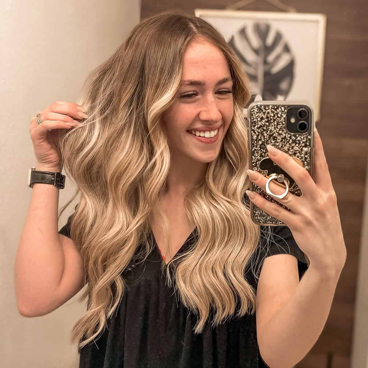 Smiling woman with long, wavy blonde hair and darker roots taking a mirror selfie while holding a section of her layered hair.