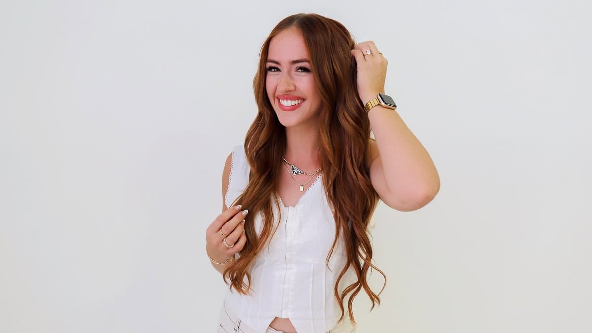Smiling woman with long, wavy auburn hair wearing a white sleeveless top and gently touching her hair against a light background.