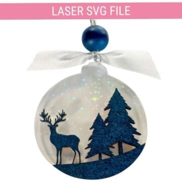 Laser-cut winter ornament featuring a deer and pine trees with a sparkly background, blue bead, and white ribbon.
