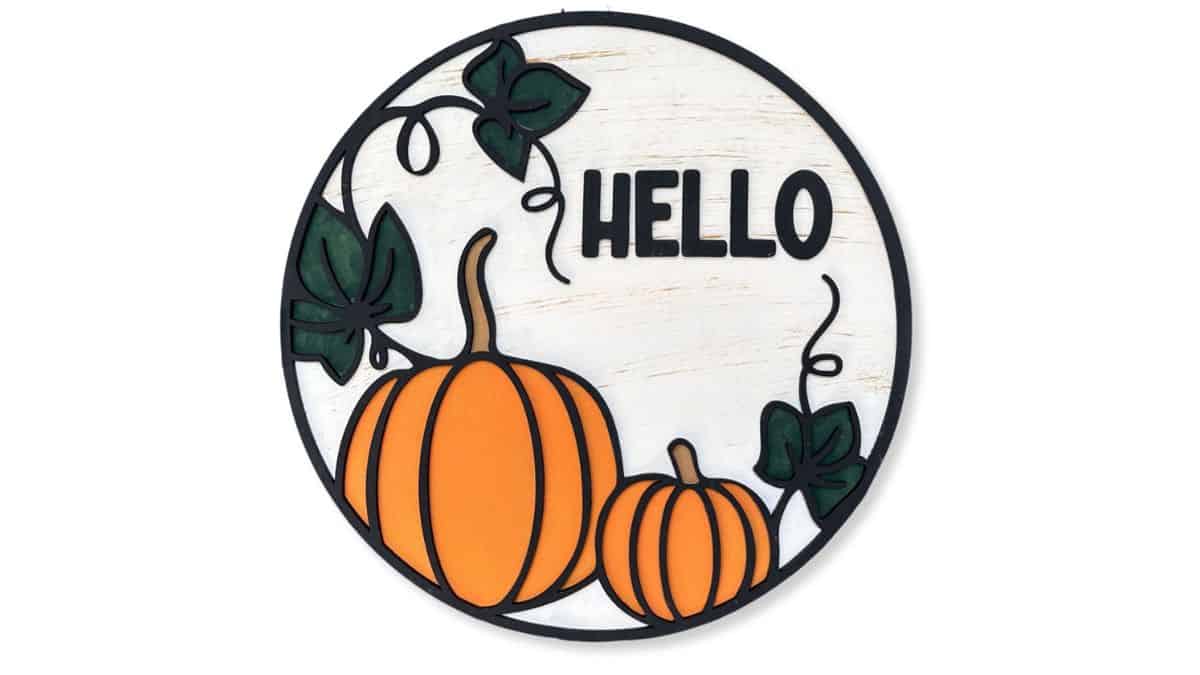 Round fall door hanger with two orange pumpkins, green leaves, and black text that reads "HELLO" on a white rustic background.