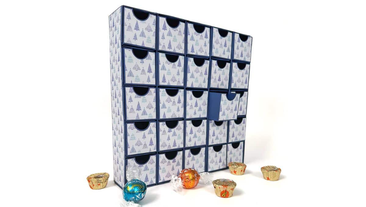 A handmade advent calendar box with multiple small drawers, decorated with a winter tree pattern, filled with chocolates and treats, with some candies scattered around.