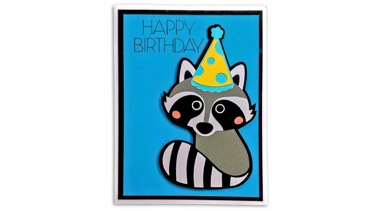 A handmade birthday card featuring a cute raccoon wearing a yellow polka-dot party hat on a bright blue background with the words "Happy Birthday" at the top.