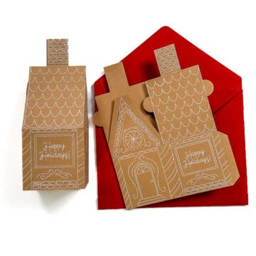 Handmade gingerbread house–shaped holiday cards with white designs and red envelopes, featuring “Happy Holidays” text.