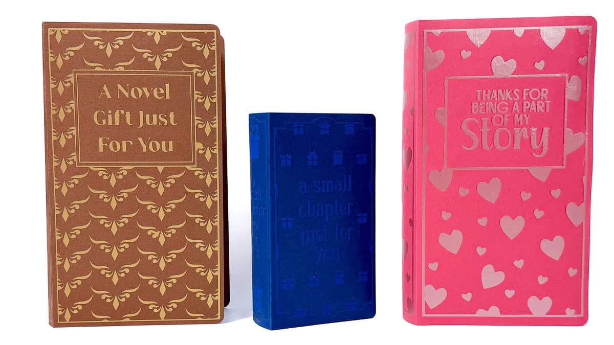 Three decorative book-shaped gift boxes in brown, blue, and pink, with phrases and patterns including hearts and ornate designs, displayed upright on a white background.